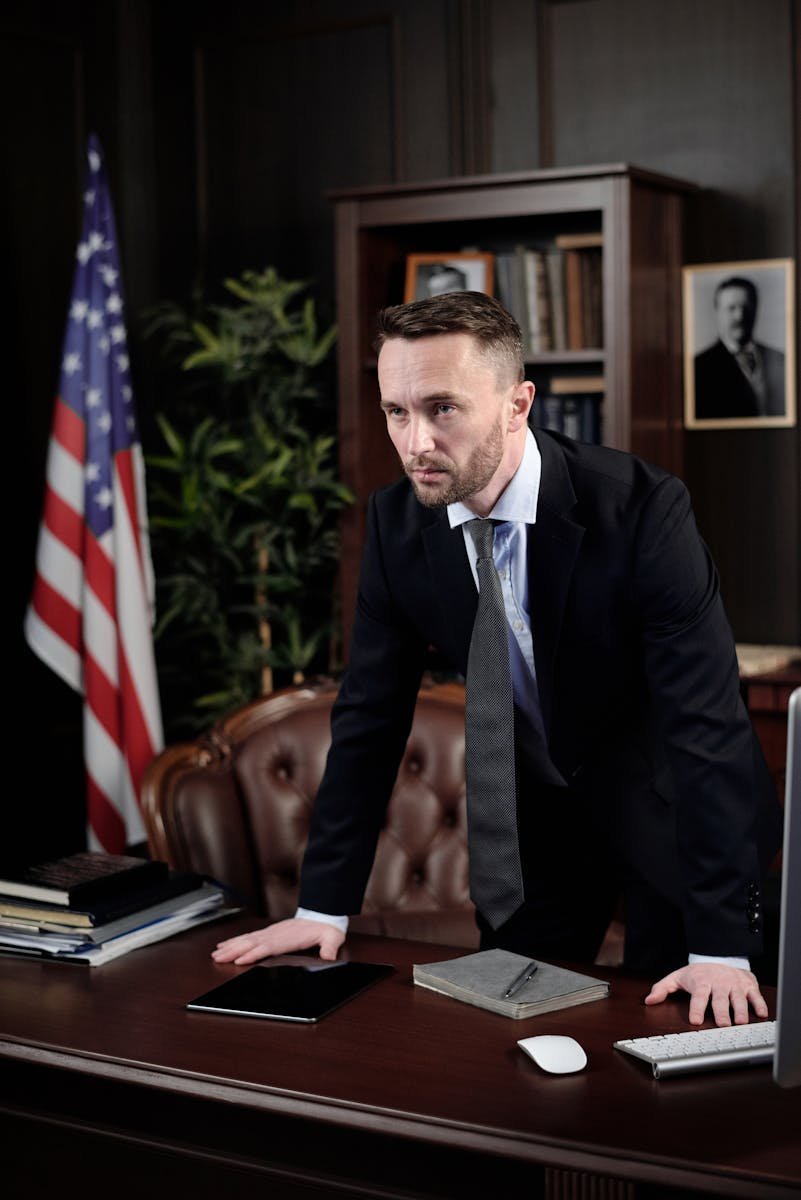 A confident man in a suit standing by a desk with an American flag in the background.